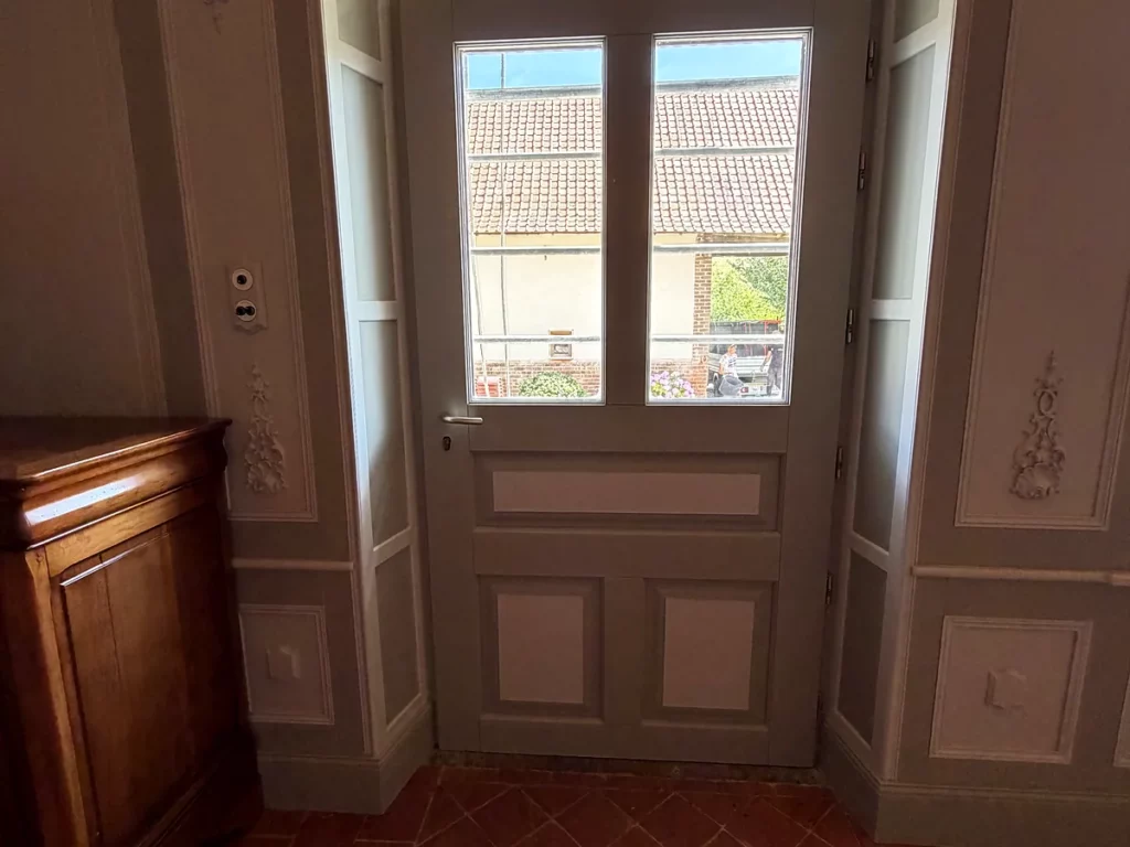 Entrée aux boiseries peintes avec panneaux moulurés et ornements sculptés, autour d'une porte vitrée en bois repeinte.