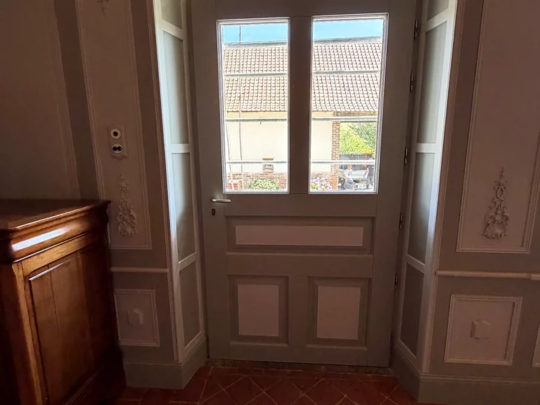 Entrée aux boiseries peintes avec panneaux moulurés et ornements sculptés, autour d'une porte vitrée en bois repeinte.