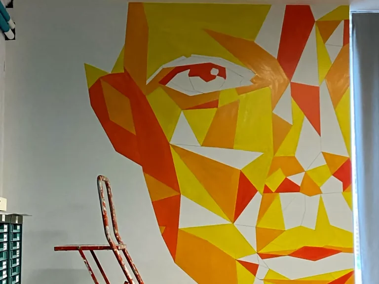 Fresque murale géométrique représentant un visage en aplats jaunes et orange, peinte à la main sur un mur de bureau.