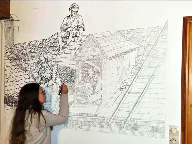 Peintre en décor traçant au feutre une fresque murale au trait représentant des couvreurs au travail sur un toit.