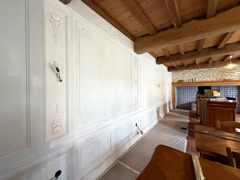 Long mur de lambris peint en blanc à moulures et ornements sculptés, sous un plafond à poutres apparentes en bois.