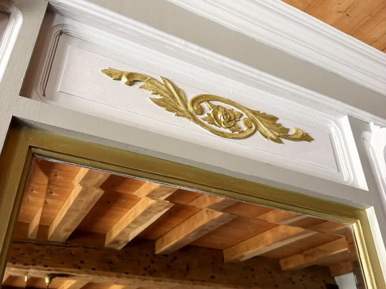 Détail d'un ornement doré à la feuille d'or sur une corniche de boiserie peinte en blanc, sous un plafond à solives.