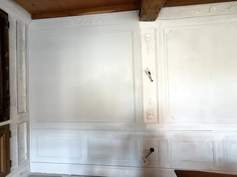 Mur de lambris peint en blanc cassé, panneaux moulurés et ornements en relief, avec applique murale en attente de finition.