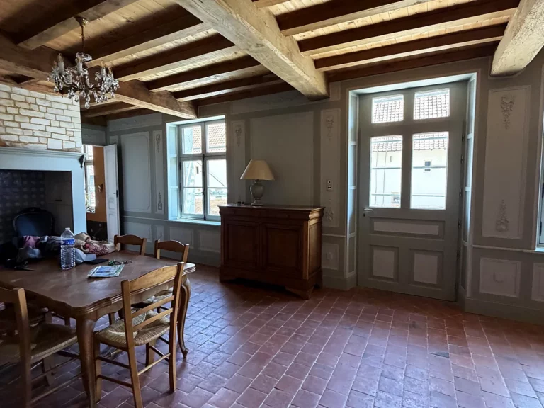 Salle à manger aux murs à panneaux peints gris et porte-fenêtre en boiserie repeinte, sous un plafond à poutres apparentes.