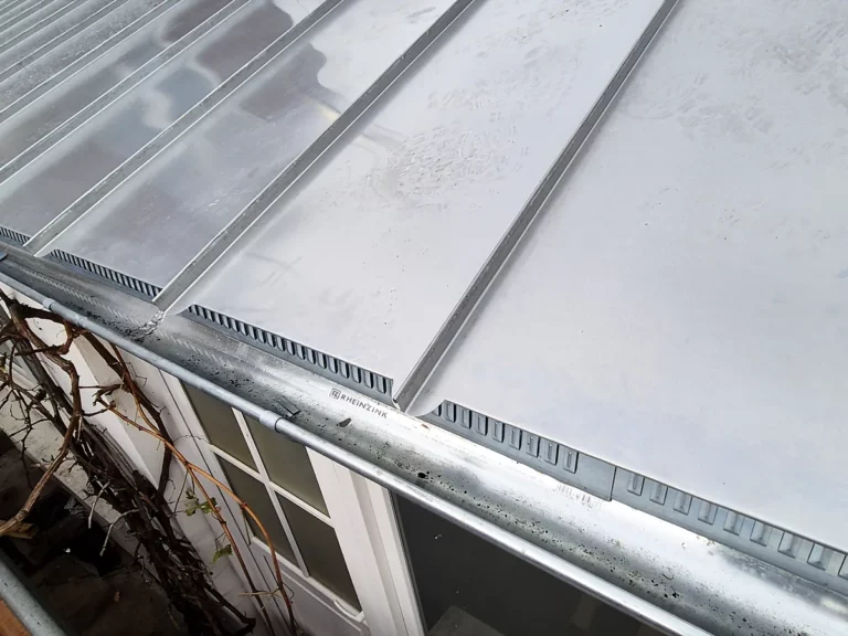 Rive et gouttière en zinc longeant une couverture à joint debout, au-dessus d'une fenêtre ancienne.
