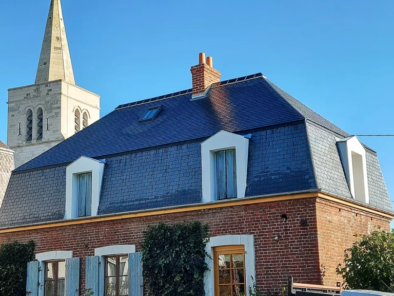 Toiture entièrement refaite en ardoise naturelle sur une maison en brique à lucarnes, avec le clocher du village derrière.